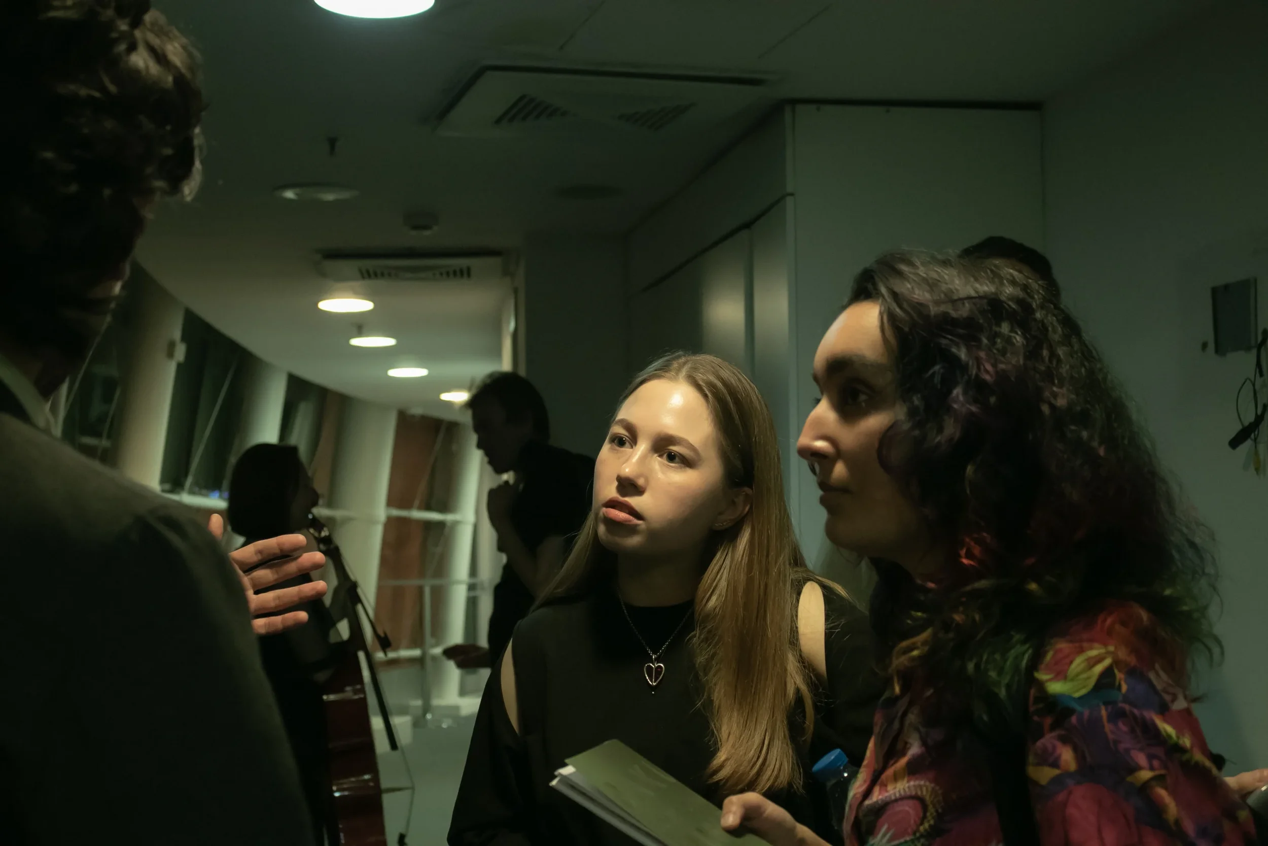Two women in conversation, one holding a notebook, in a dimly lit indoor setting with a corridor and other people in background.