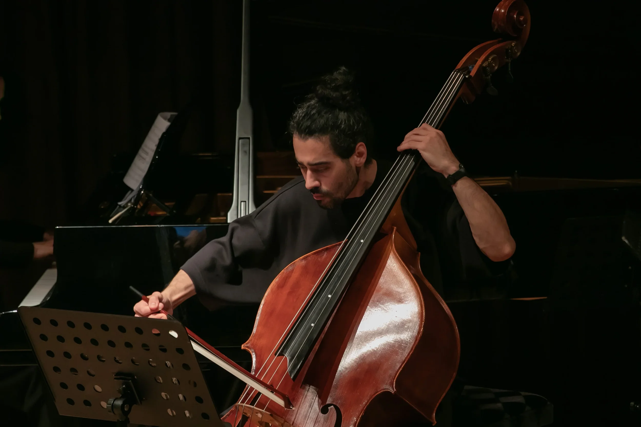 A man playing a double bass in a music performance, with a piano and sheet music visible in the background.