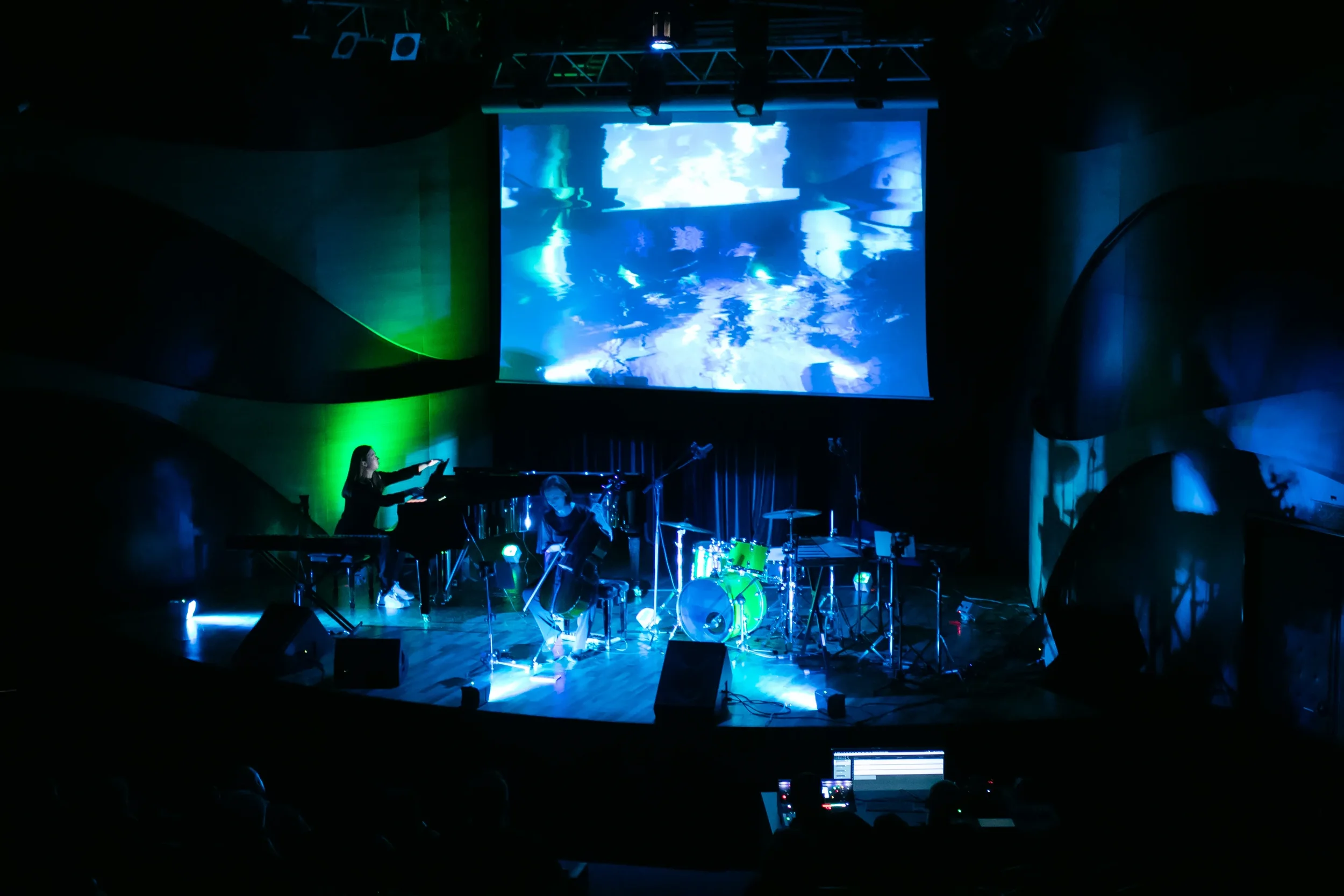 An indoor stage with musicians, including a keyboardist, string player, and drummer, illuminated by blue and green lights, with a large screen displaying abstract visuals overhead.