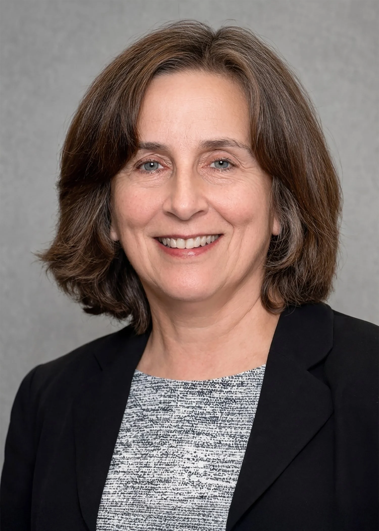 A woman with brunette hair smiling, wearing a black jacket and a gray textured blouse.
