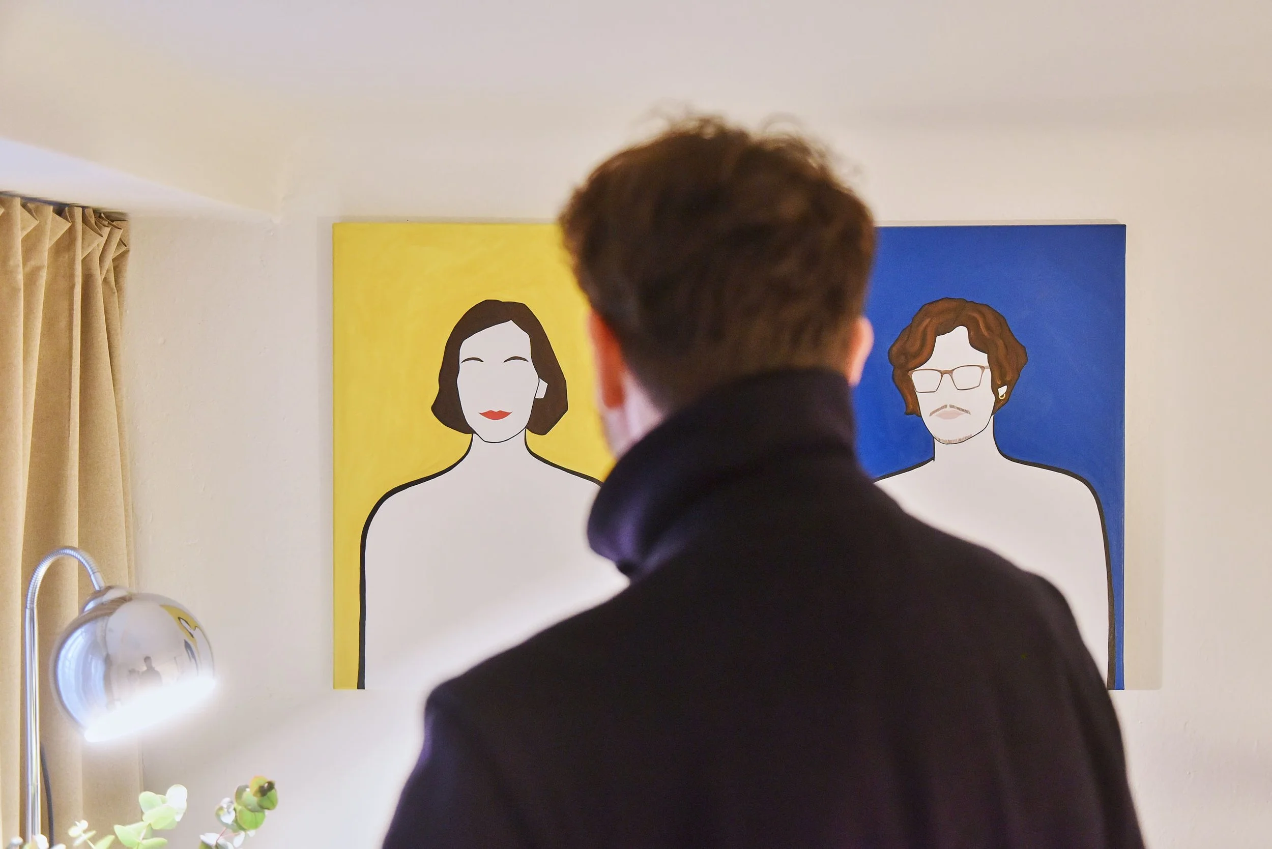 Uomo con capelli castani corti, visto di spalle, che guarda due quadri raffiguranti persone stilizzate, uno con sfondo giallo e l'altro con sfondo blu, in un ambiente domestico con lampada, piante e tende beige.