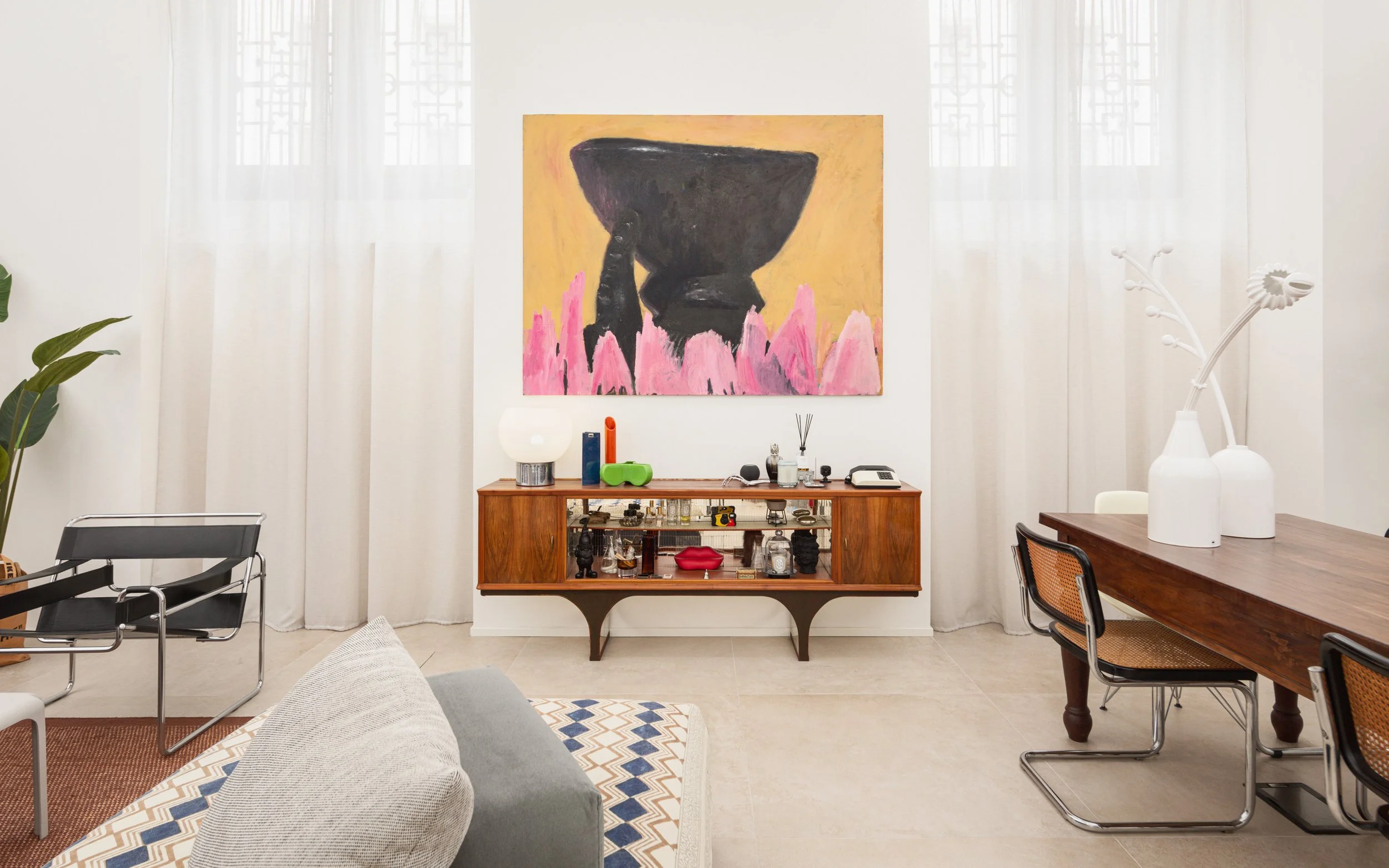 Interno di un soggiorno con arredi moderni e un mobile in legno, grande quadro astratto sulla parete, divano con cuscino, tavolo in legno con sedie, vasi biani con fiori decorativi e oggetti vari.