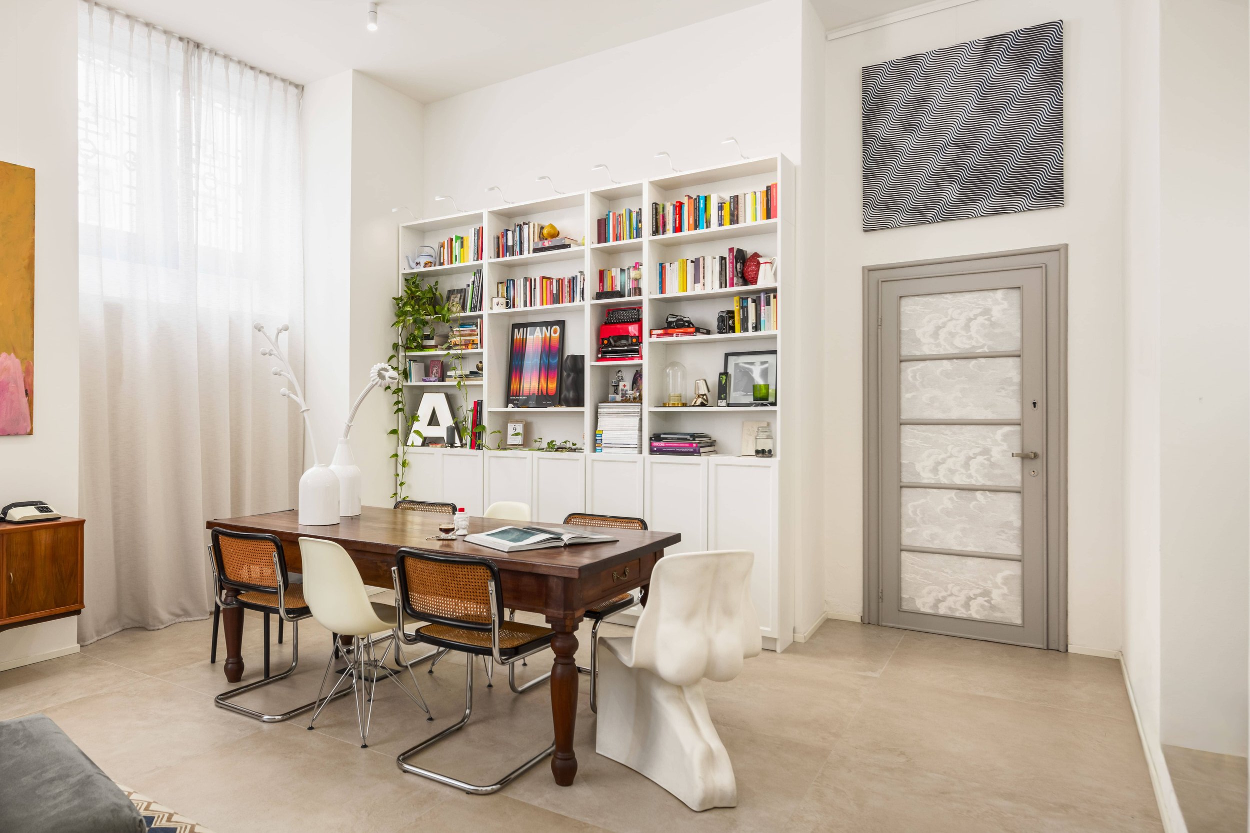 Sala da pranzo con tavolo in legno, sedie di vari stili, libreria bianca piena di libri e decorazioni, pareti bianche, porta grigia, finestre con tende leggere biancastre, decorazioni moderne e arredi minimalisti.