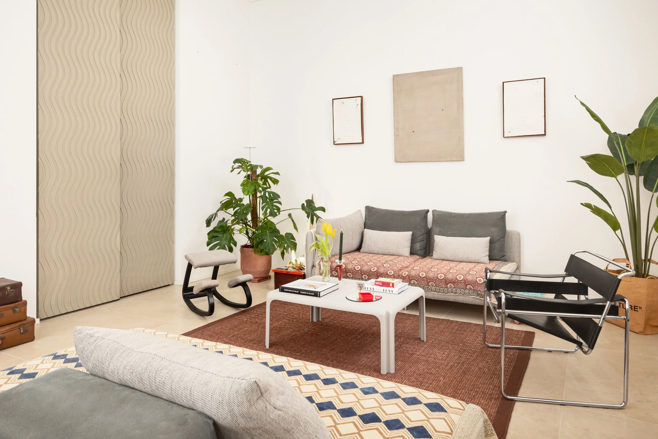 Soggiorno con divano grigio, tappeto marrone, tavolino bianco con libri e oggetti, due piante grandi, una sedia moderna in metallo e tessuto nera, quadri minimalisti alle pareti, ambientazione accogliente e moderna.