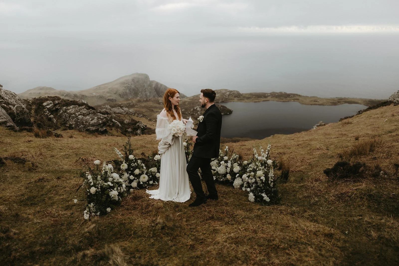 Ein Brautpaar bei einer Hochzeit im Freien vor einer ruhigen Seenlandschaft mit Bergen im Hintergrund, umgeben von weißen Blumen.