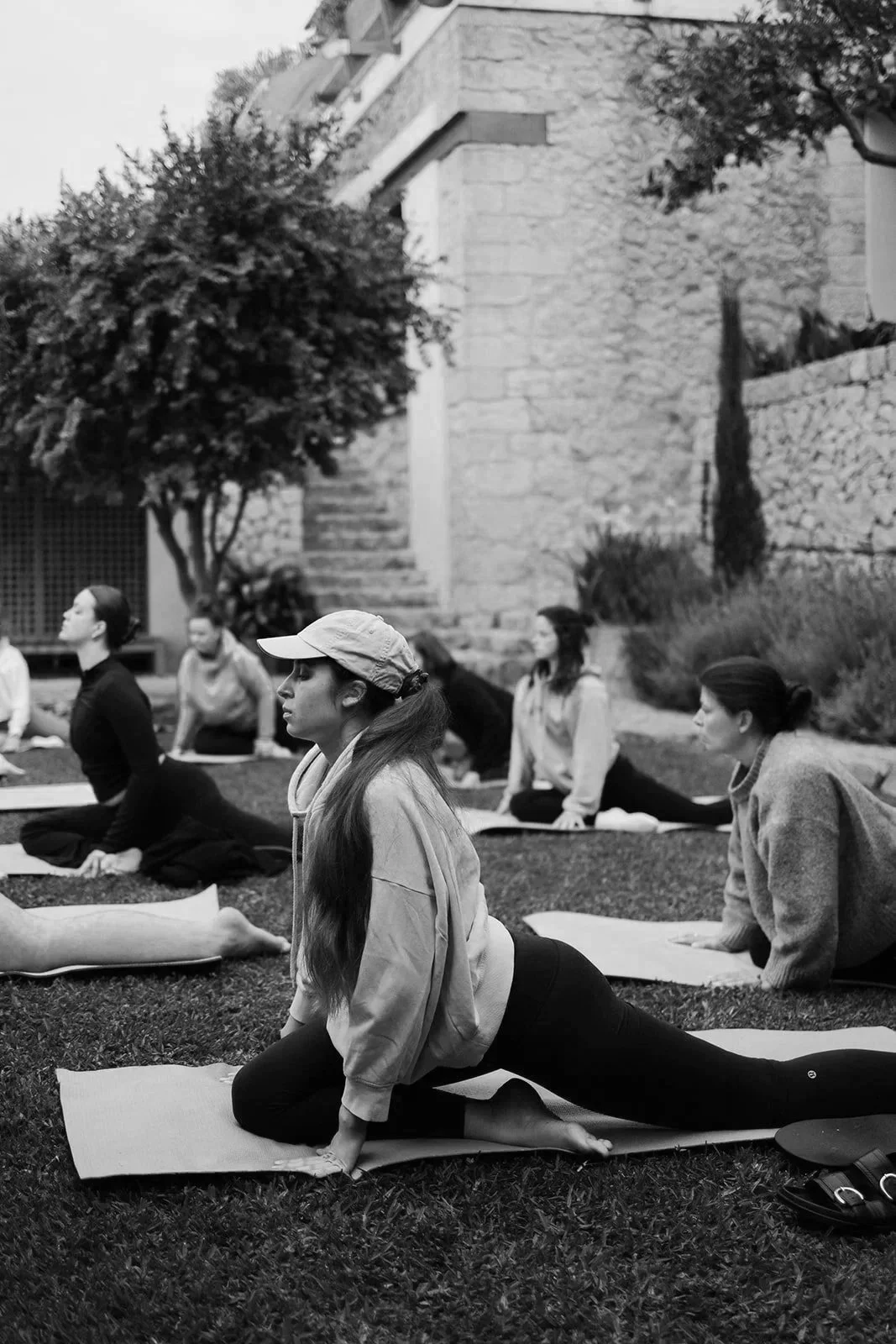 Gruppe von Frauen beim Yoga im Freien, in einer entspannten Haltung auf Yogamatten, umgeben von Bäumen und einer Steinmauer.