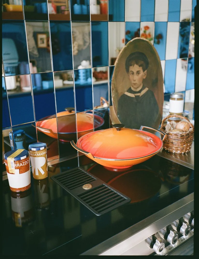 Cucina con piani cottura, barattoli di alimenti, una ciotola con coperchio, un quadro con un ritratto di un bambino, uno scaffale con oggetti decorativi e piante, sullo sfondo piastrelle blu e arancioni.