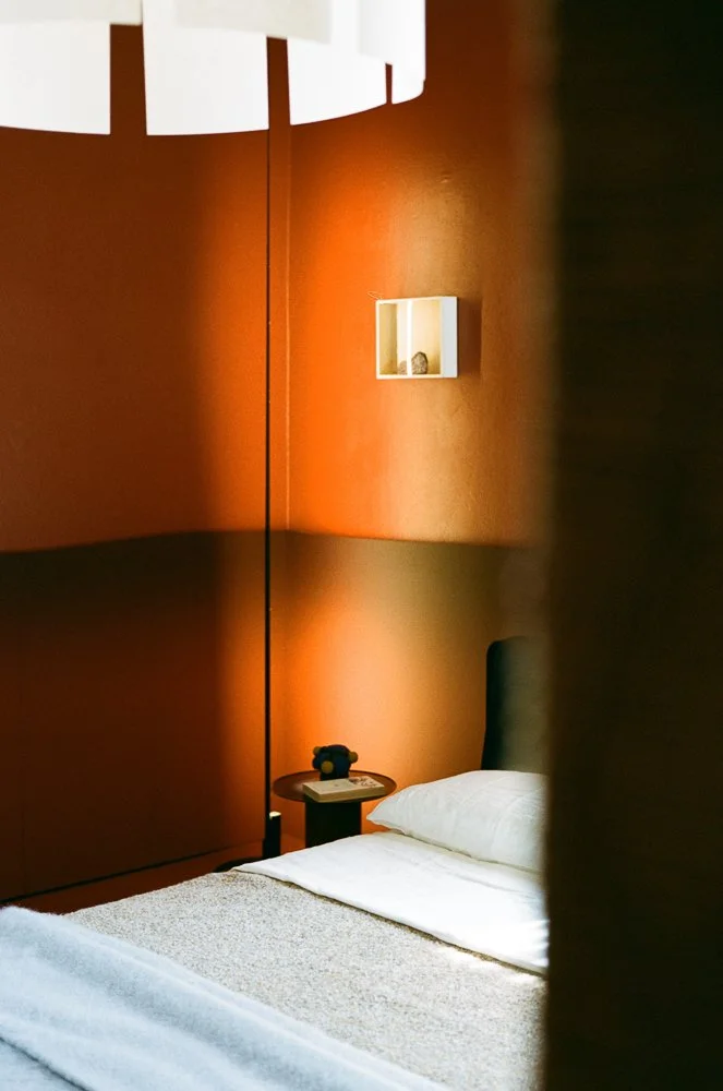Camera da letto con pareti arancioni, letto con lenzuola bianche, comodino con telefono e animale di pezza, mensola decorativa sulla parete.