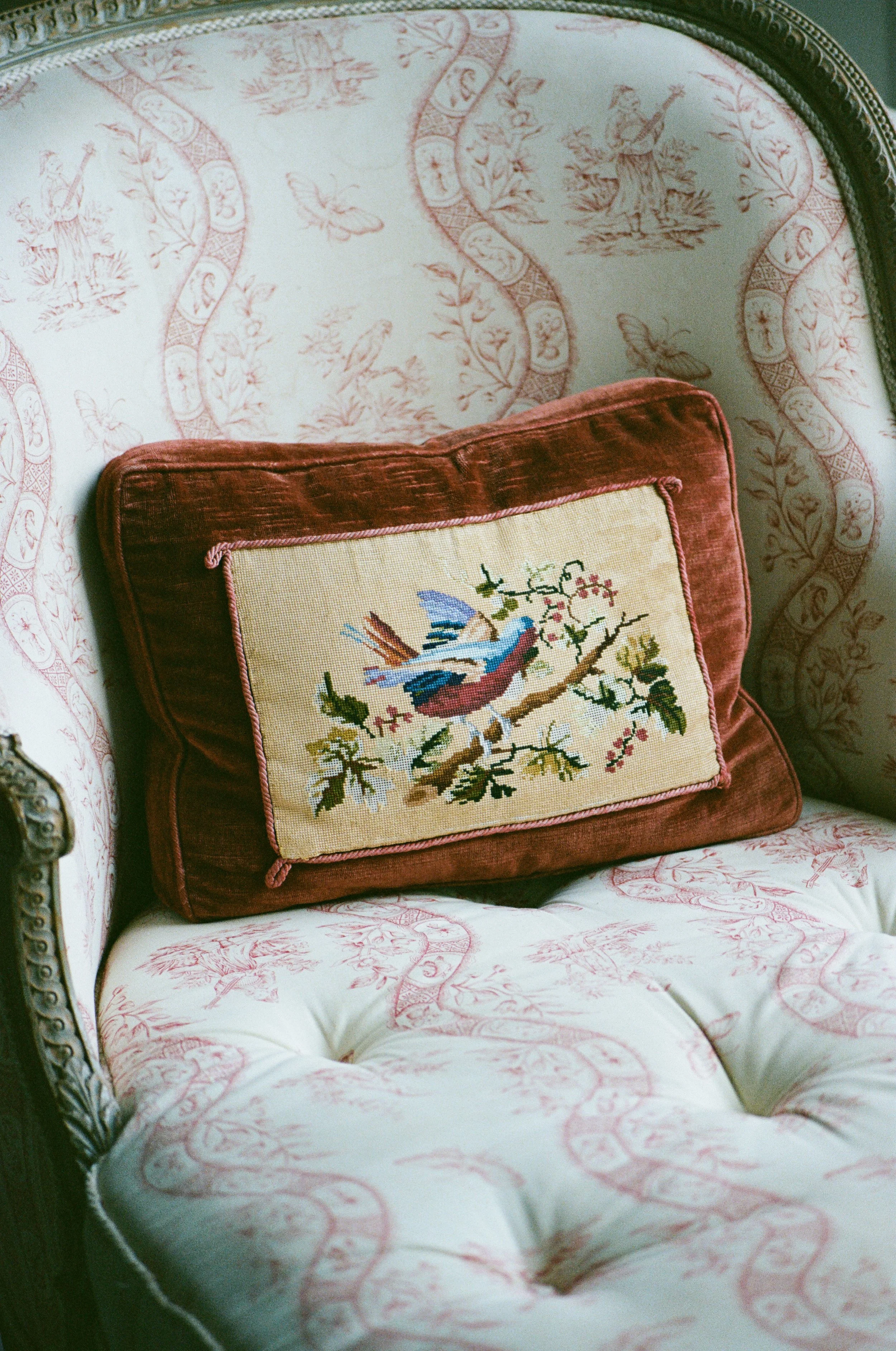 Cuscino ricamato con un uccello su un ramo, appoggiato su una poltrona con tessuto ricamato a fiori e foglie.