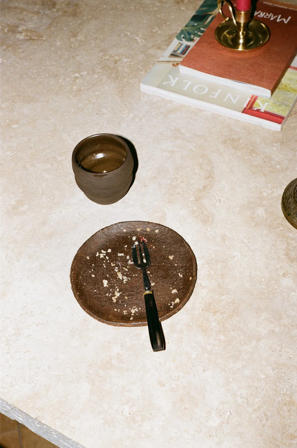 Piattino sporco con forchetta e briciole, bicchiere vuoto, libro e fumaiolo acceso, tutto su un tavolo.