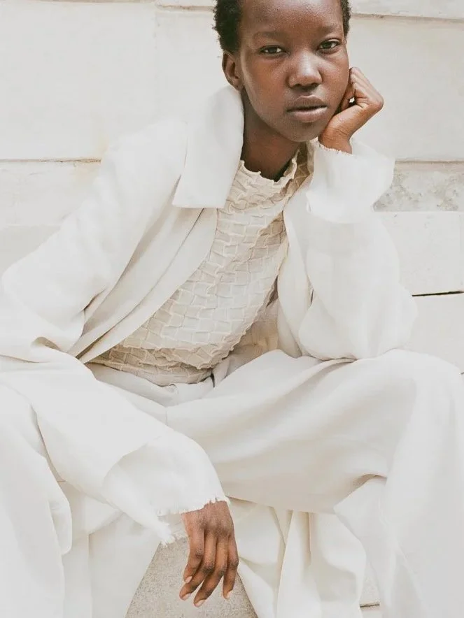 Una donna nera seduta contro un muro bianco, indossa abiti chiari e guarda verso la fotocamera con espressione seria.