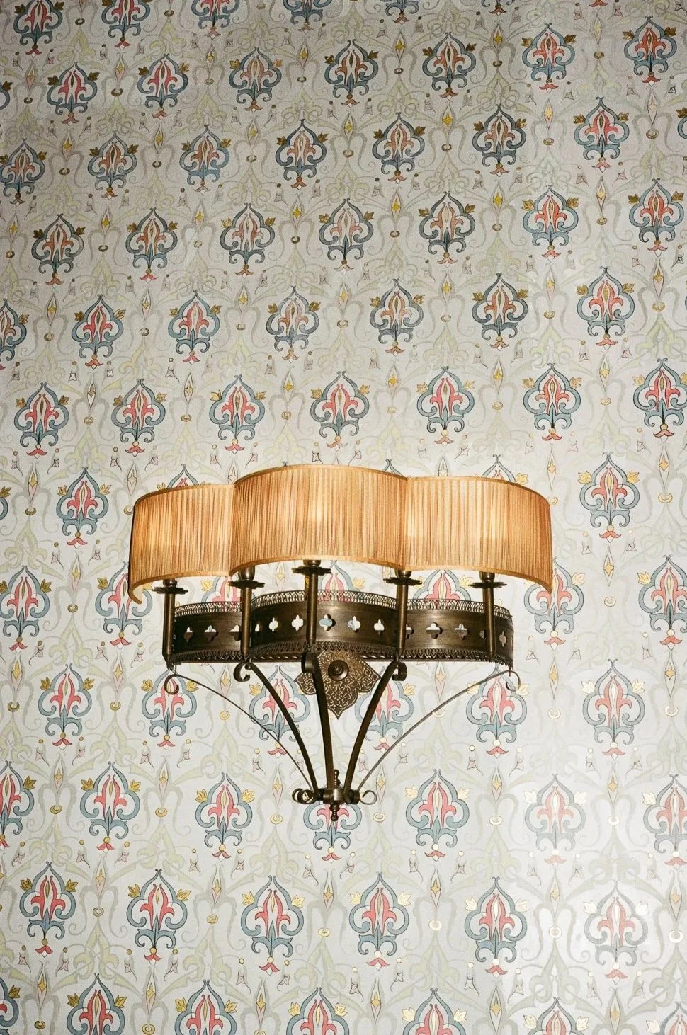 Una lampada da parete con paralume dorato e struttura in ferro nero, montata su una parete con parete decorativa con motivo floreale in tonalità pastello