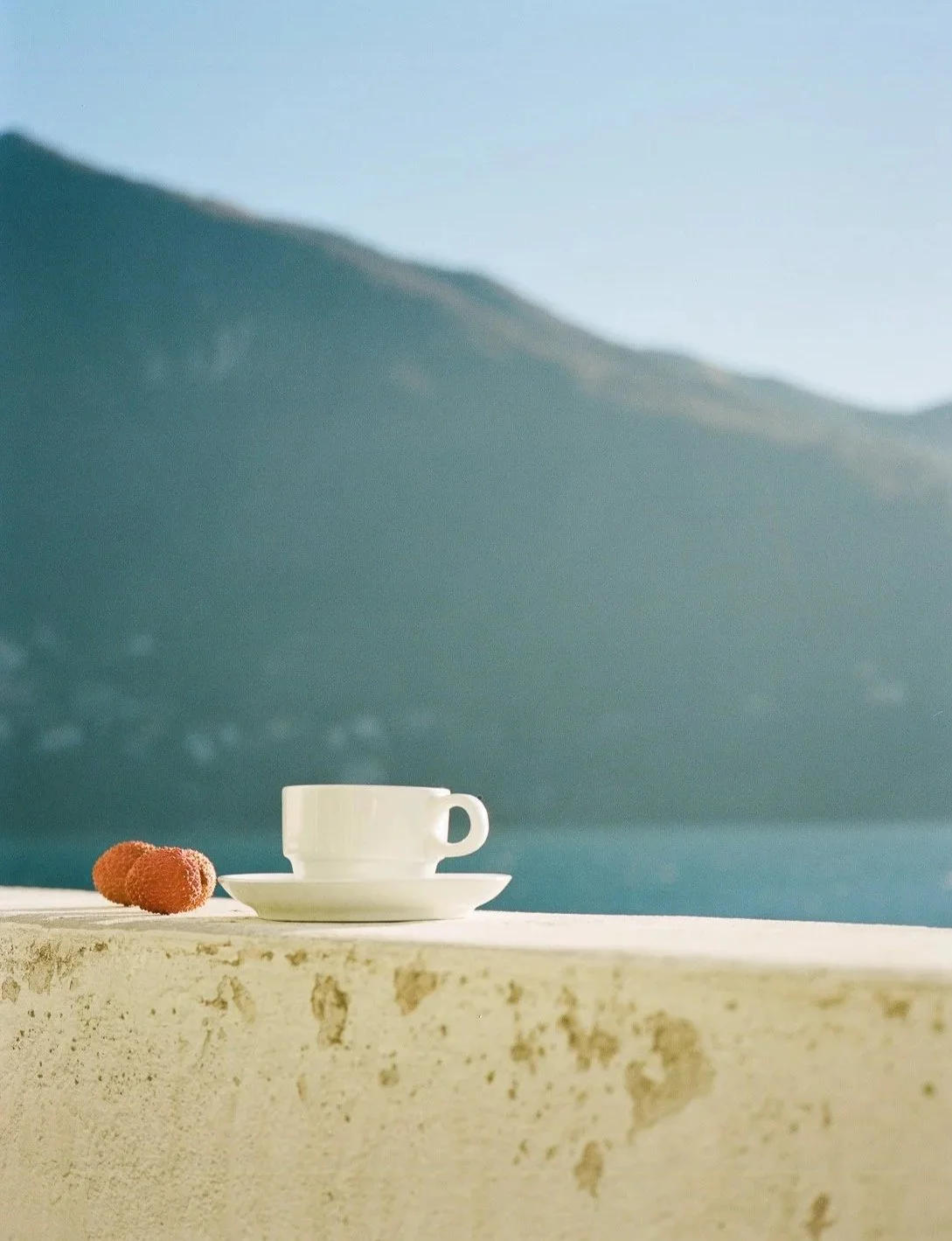 Tazza bianca su una piattina su una superficie beige, con due frutti rossi accanto, sullo sfondo montagna e cielo azzurro