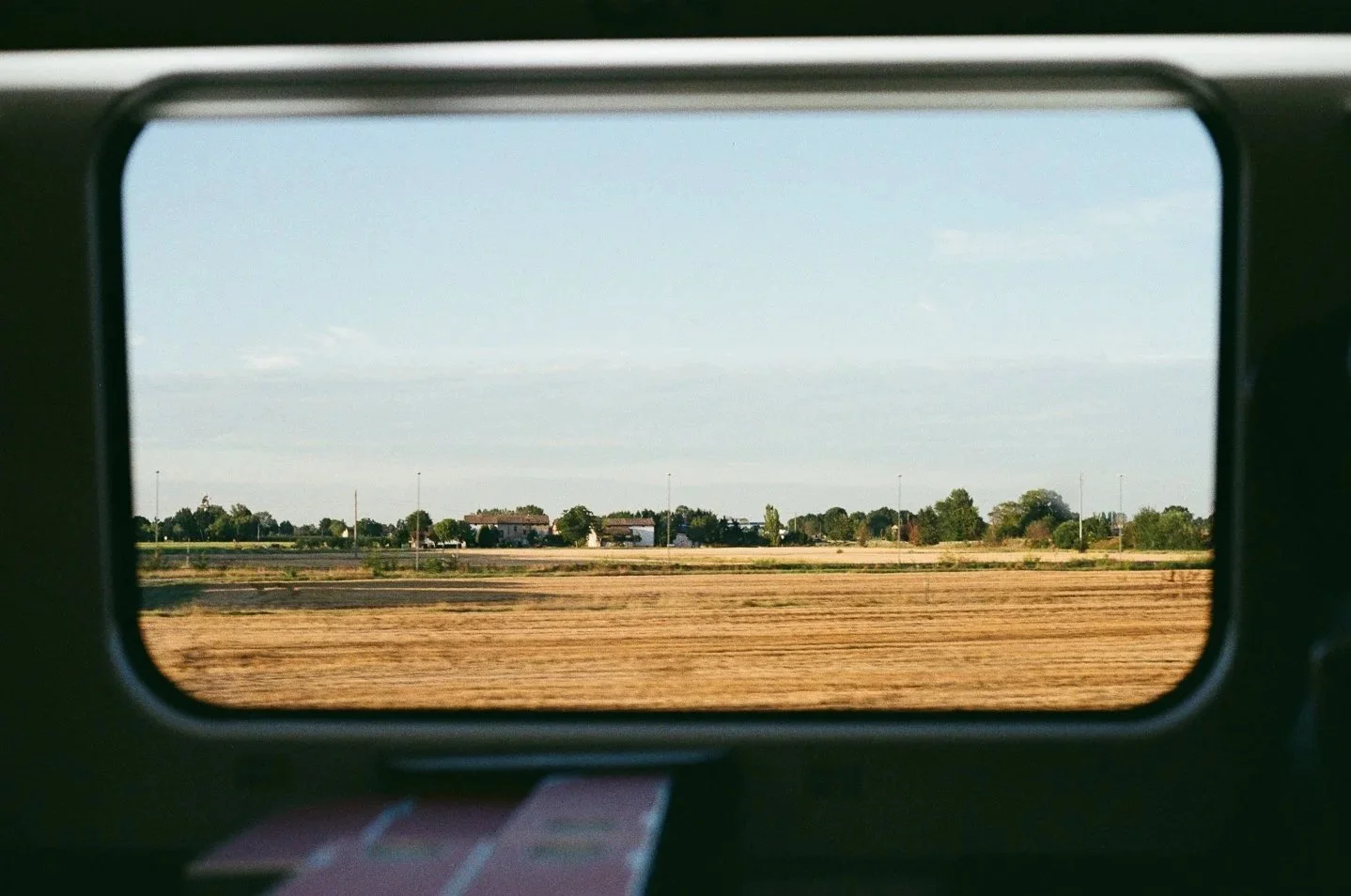Vista di un paesaggio rurale attraverso il finestrino di un treno che corre su campi coltivati e alcune case distanti.
