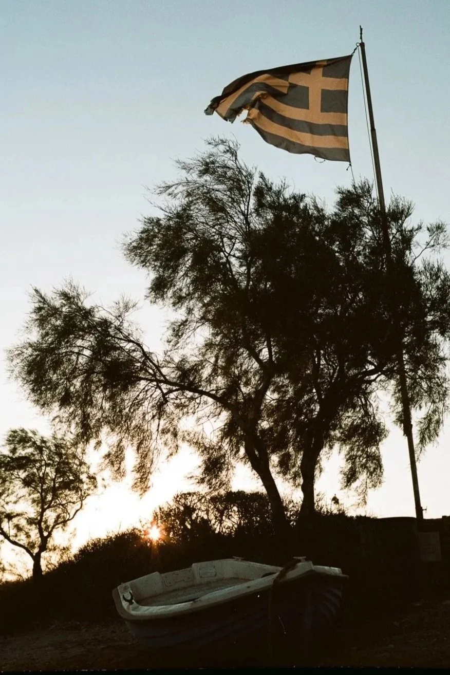 Albero con bandiera greca stilizzata che sventola al vento, al tramonto, con barca nel primo piano.