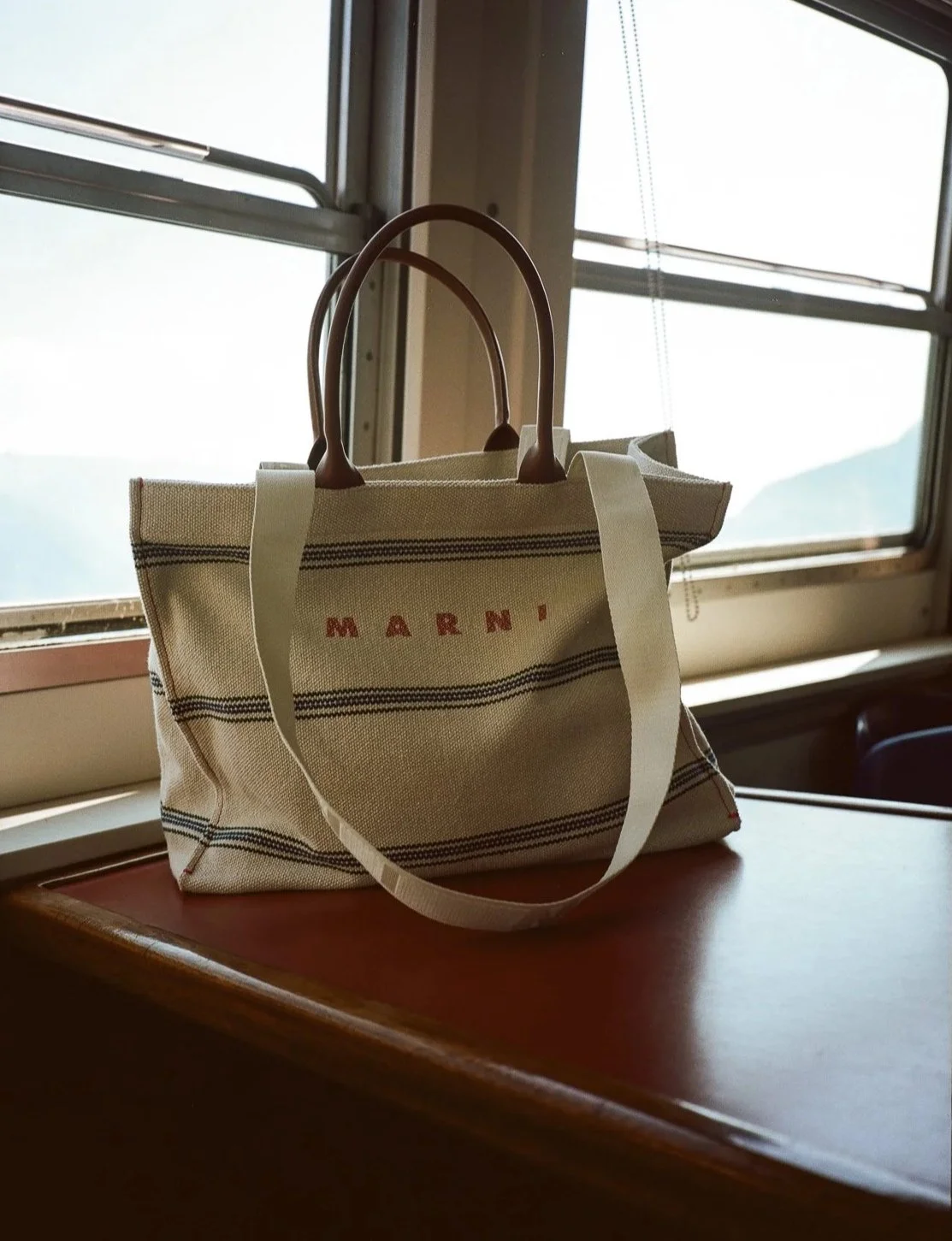 Borsa a mano di tela con doppie maniglie di pelle, posizionata su un tavolo vicino a una finestra con vista di montagne.