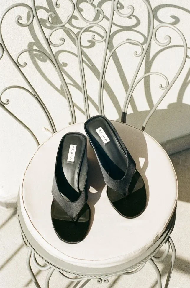 Sandali neri con tomaia aperta e logo "ALAIIA" posizionati su una sedia bianca con schienale decorato in metallo.