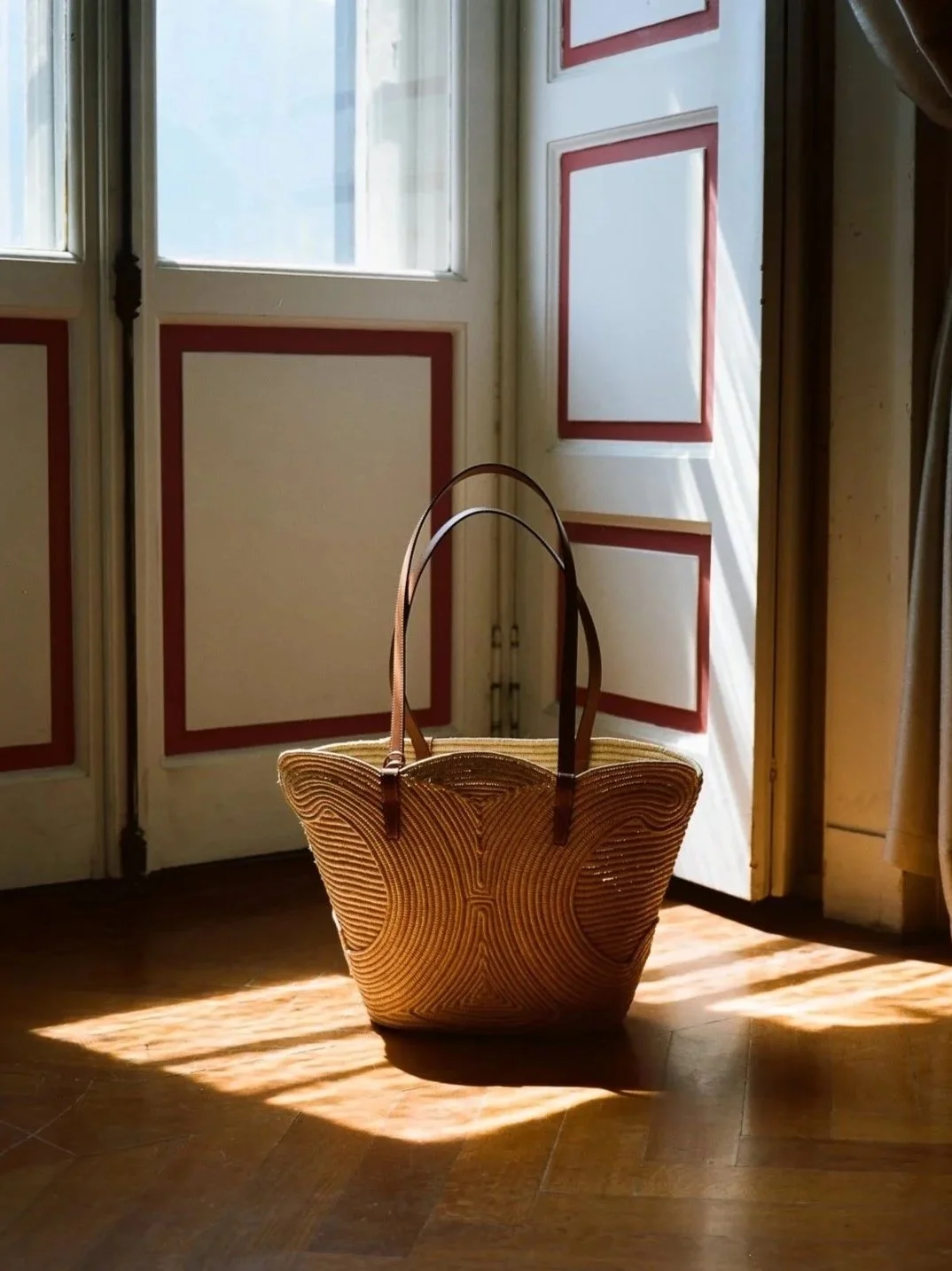 Una borsa di paglia con manici di pelle, collocata a terra vicino a una porta finestra luminosa.