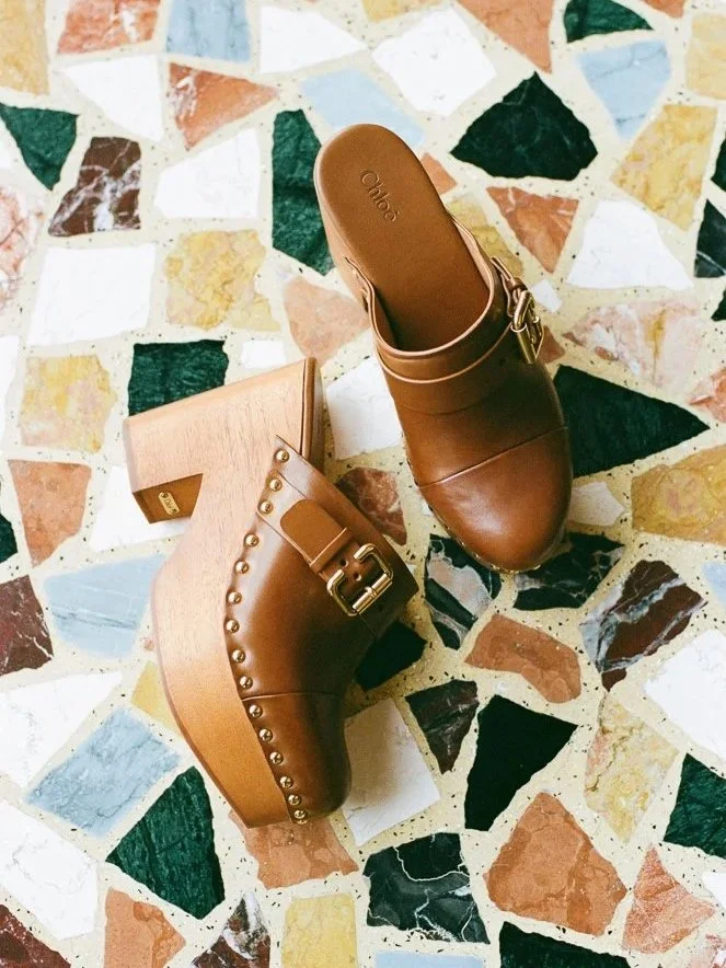 Scarpe in pelle marrone con tacco a blocco e dettagli di borchie metalliche, poggiate su un pavimento in mosaico colorato.