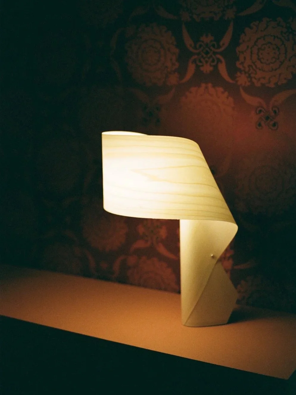 Lampada da tavolo accesa su una superficie con carta o legno chiaro, sfondo con carta da parati rossa scura con motivi decorativi