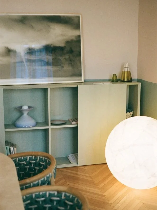 Interno con scaffale bianco, lampada di carta luminosa, sedie con rivestimento particolare, quadro con cielo nuvoloso.