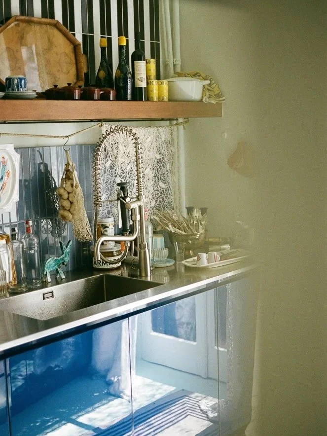 Angolo di una cucina con lavabo in acciaio, sopra uno scaffale con bottiglie di vino, barattoli e altri oggetti. Sul lavandino ci sono piatti, tazze e utensili vari, con uno sfondo di tende ricamate e una porta finestra che lascia entrare luce natura