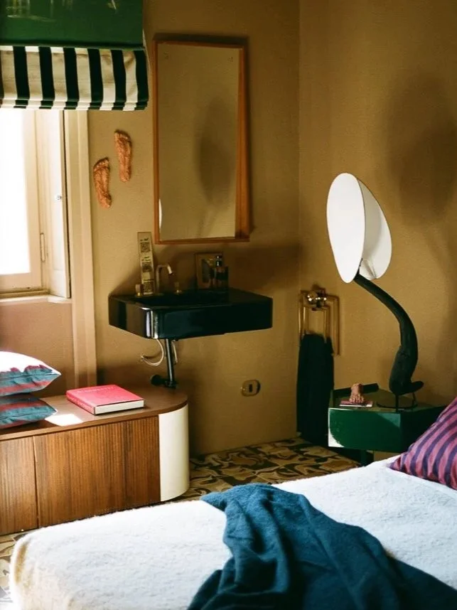 Camera da letto con letto coperto da una coperta beige, ci sono tessuti blu e a strisce, mobile in legno con libri e cuscini, specchio e lavabo nero sopra un mobile, lampada moderna accanto al letto.