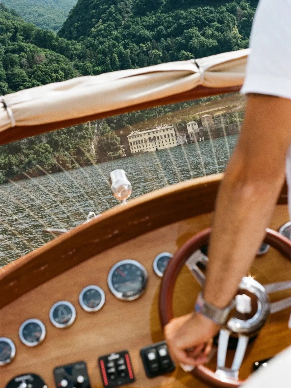 Vista di una barca che naviga su un lago, con uno sfondo di colline verdi e un edificio bianco sulla riva.