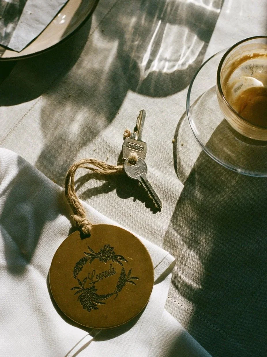 Chiavi con portachiavi dorato, tazza di caffè, bicchiere di acqua, e un piattino con un bicchierino di liquore su una tovaglia chiara, con ombre e riflessi di luce.