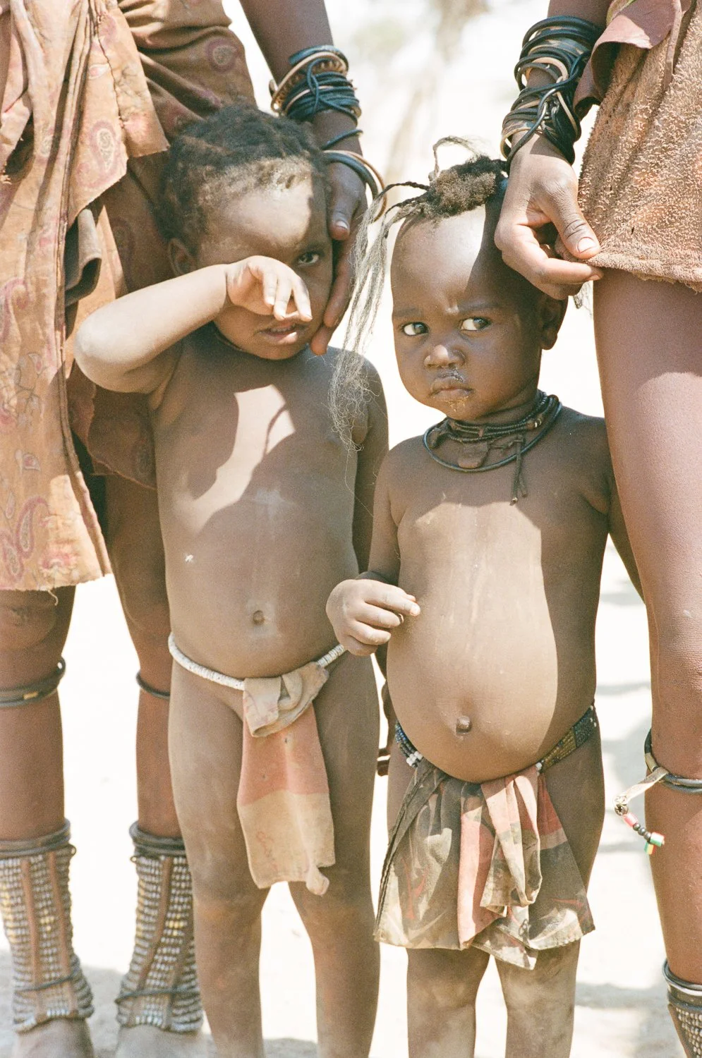 Due bambini nudi con collane e abiti tradizionali, uno con il braccio sollevato e l'altro che guarda verso la fotocamera, circondati da adulti.