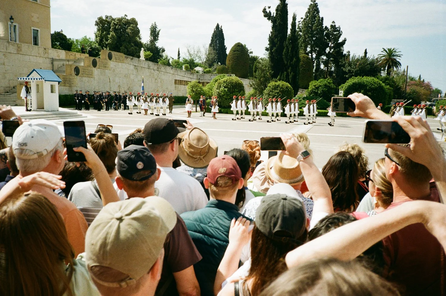 Più persone assistono a una parata militare all'aperto, alcune scattando foto con il telefono, con un gruppo di soldati vestiti con abiti tradizionali e uniformi militari che sfilano davanti a loro, con un'area pavimentata, alberi e edifici sullo sfo