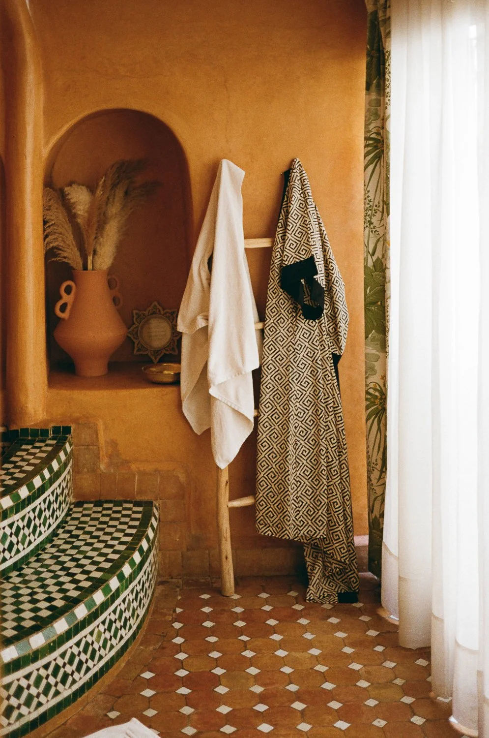Spazio interno con pareti di colore beige, scale a seconda di piastrelle verde e bianco, un'alcova con vasi e decorazioni, un appendiabiti con asciugamani e vestiti, e una tenda di tessuto bianco con motivi verdi e foglie tropicali.