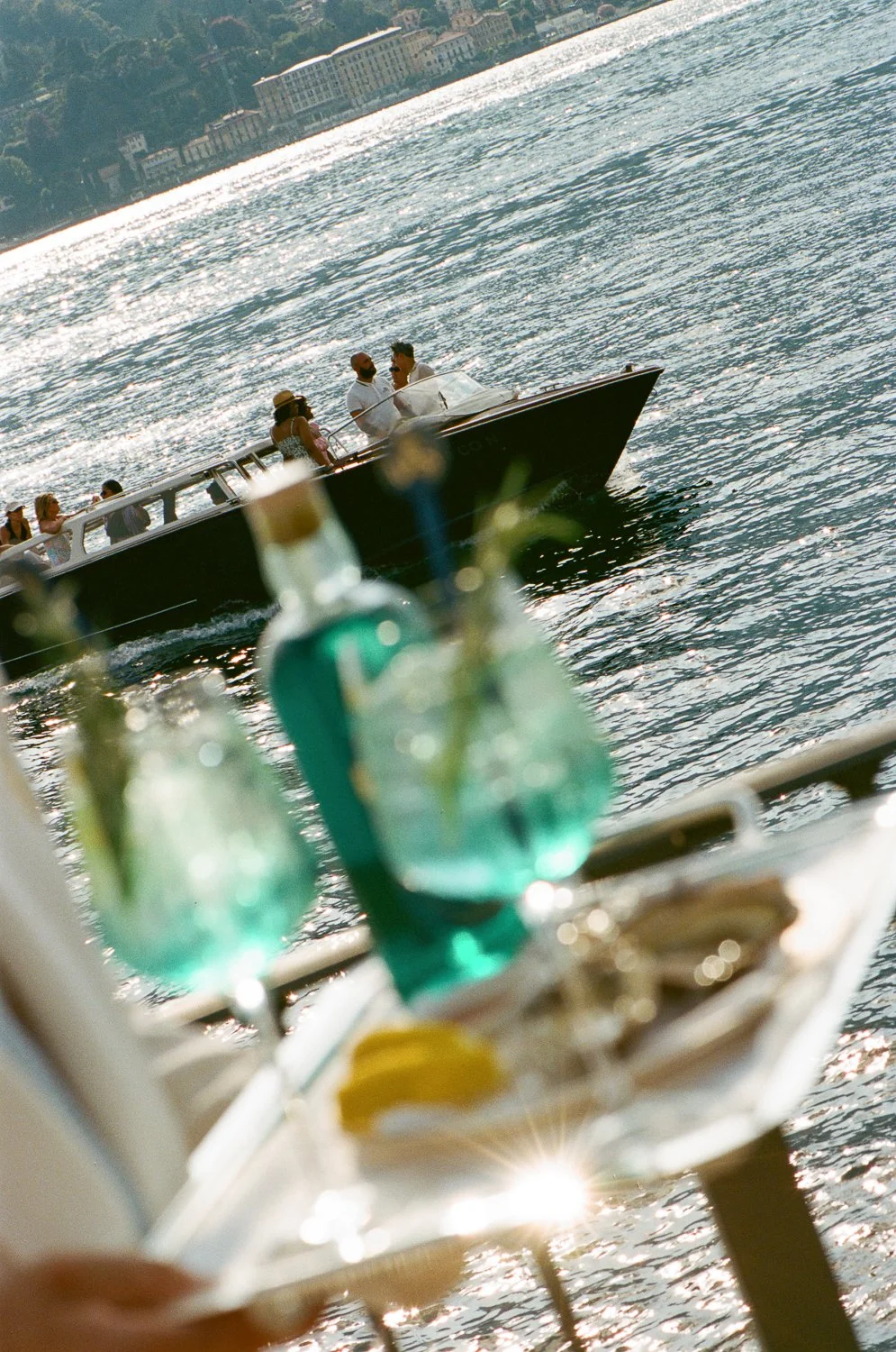 Cena con bevande, piatto di limoni, sfondo acqua con barca e persone che socializzano a bordo, sullo sfondo skyline di edifici e natura.