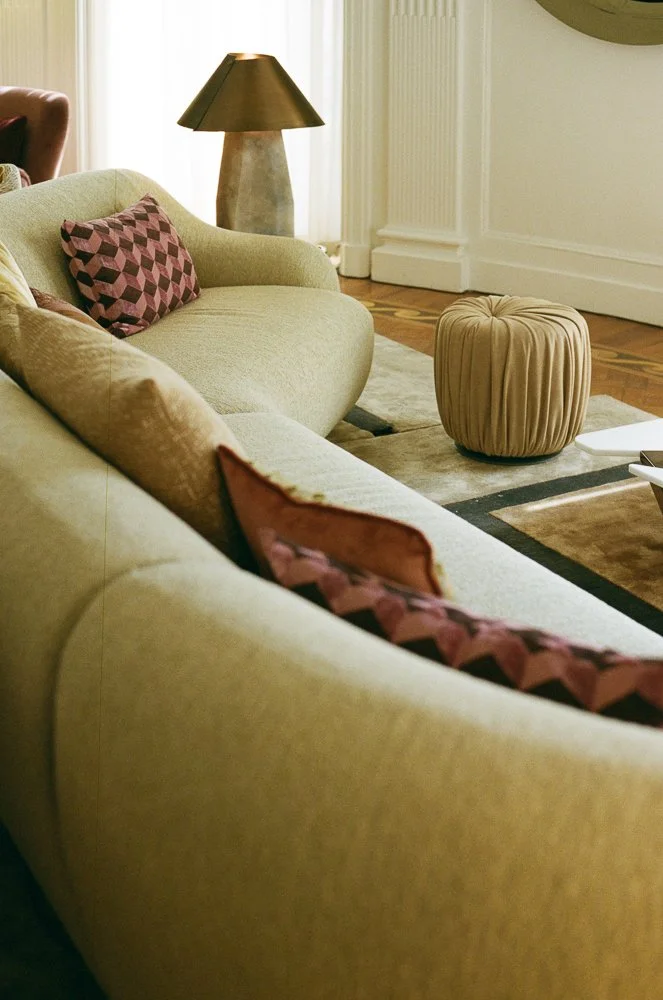 Soggiorno elegante con divani beige, cuscini decorativi con motivi geometrici rossi e neri, lampada di colore oro sulla sinistra, tavolino rotondo morbido con rivestimento beige al centro, pareti chiare e parquet in legno.