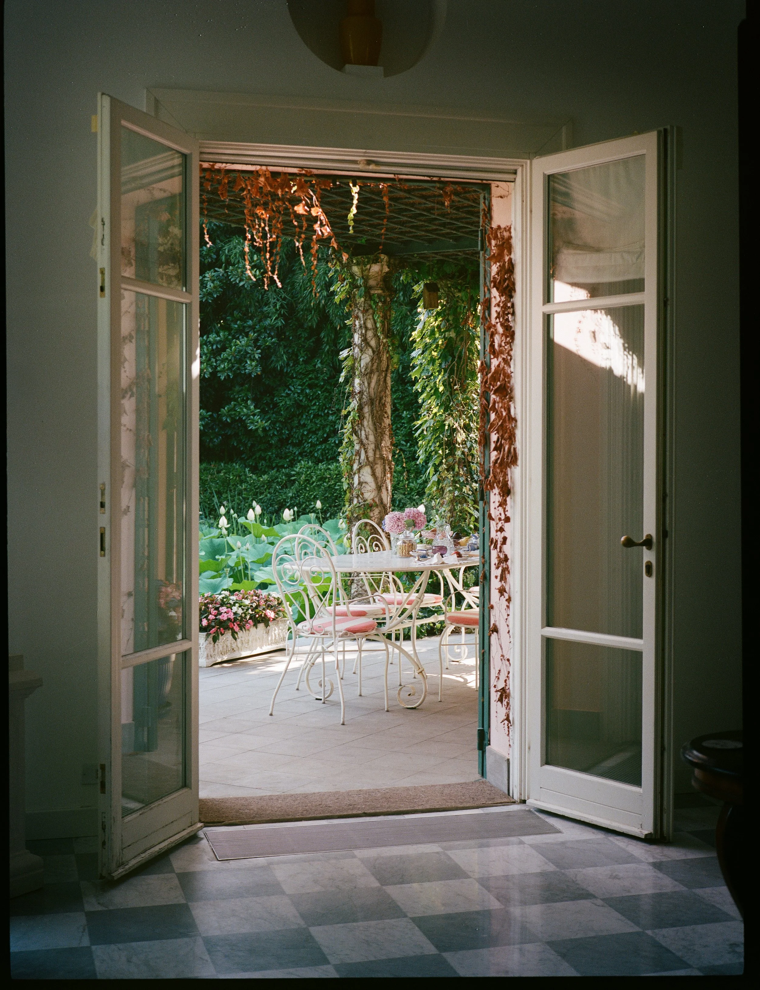 VISTA DELLA PORTA APERTA SU UN TERRAZZO O GIARDINO CON UN TAVOLO E SEDIE BIANCHI E UNA VANCHETTA CON FIORI, CIRCONDATO DA PIANTE VERDI E ALBERI.