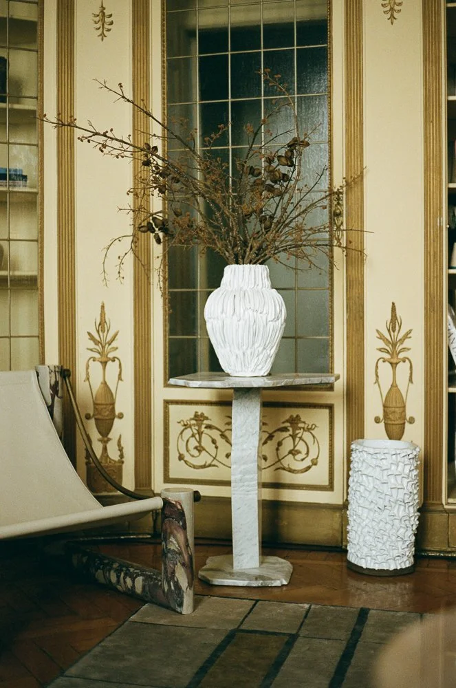 Vaso bianco con rametti secchi e foglie secche, posato su un cavalletto di marmo, in una stanza con pareti decorate dorate e un mobile con ante a vetro.