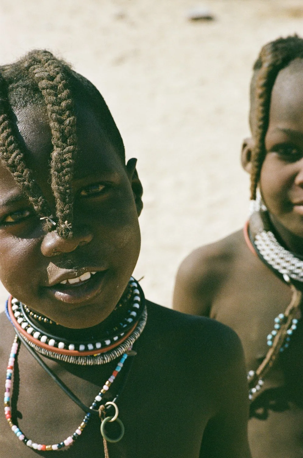 Due donne con trecce, indossano collane tradizionali di perline, scattato in un ambiente soleggiato.
