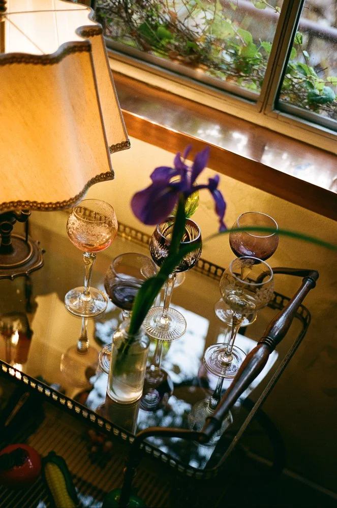 Una tavola con un vaso di vetro con un fiore viola, cinque bicchieri di vino con diversi colori, una lampada da tavolo e una finestra con vetri che mostrano foglie verdi all'esterno.