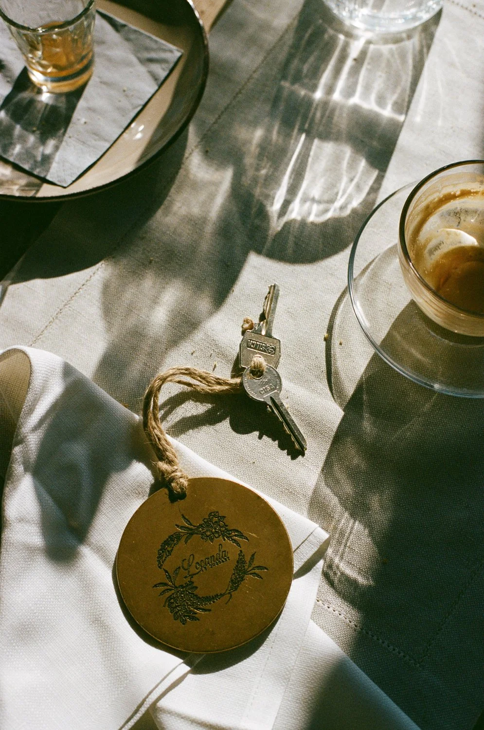 Chiavi con portachiavi dorato, tazza di caffè, bicchiere di acqua, e un piattino con un bicchierino di liquore su una tovaglia chiara, con ombre e riflessi di luce.