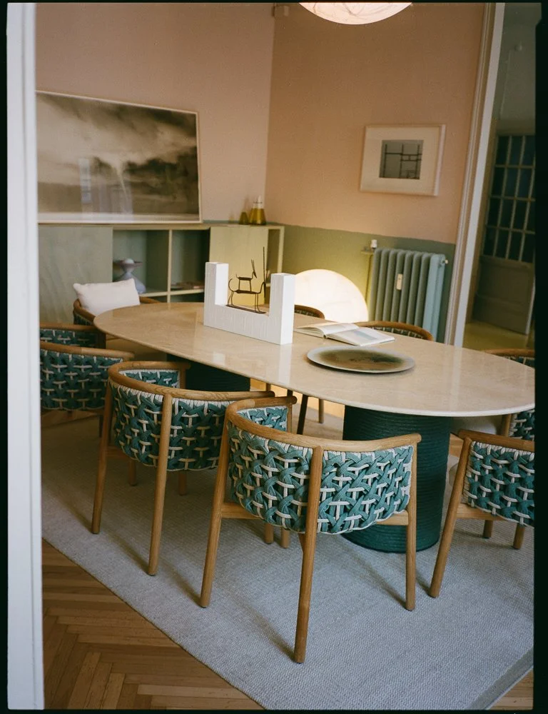 Una sala da pranzo con un grande tavolo ovale, sedie di legno con sedute intrecciate blu e beige, un grande piatto, un libro aperto e un oggetto decorativo triangolare bianco con una sedia in miniatura sopra. Sullo sfondo, una libreria bianca, un qua