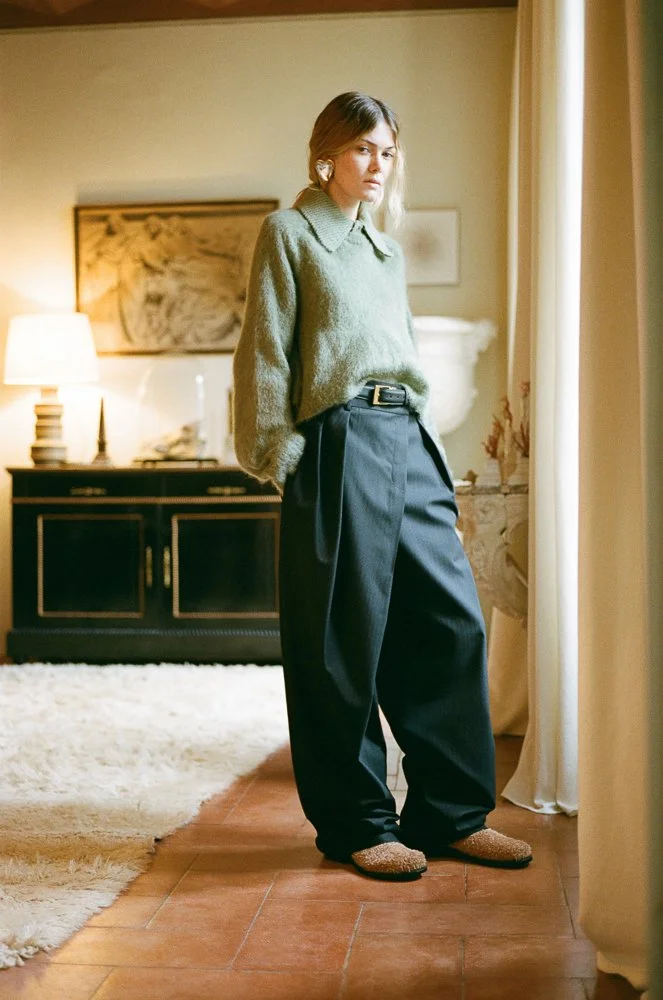 Una donna in abiti casual con un maglione grigio e pantaloni larghi neri si trova in una stanza ben arredata con una tenda, un quadro e una lampada.