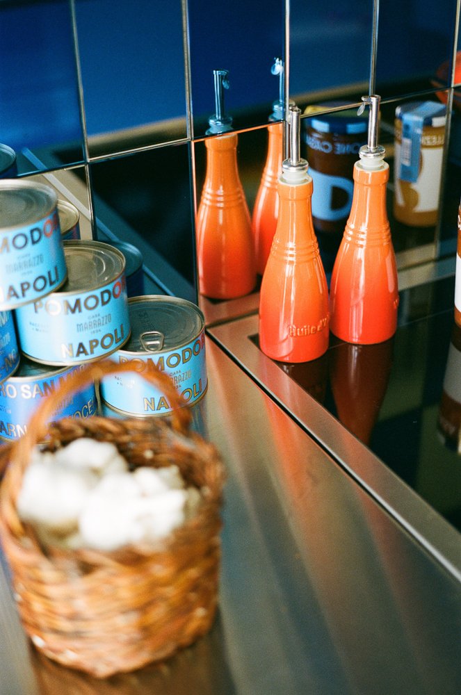 Contenitori di crema di peperoncino e scatole di conserve di pomodoro su un banco di cucina, con cappelle di pane in un cestino in primo piano.