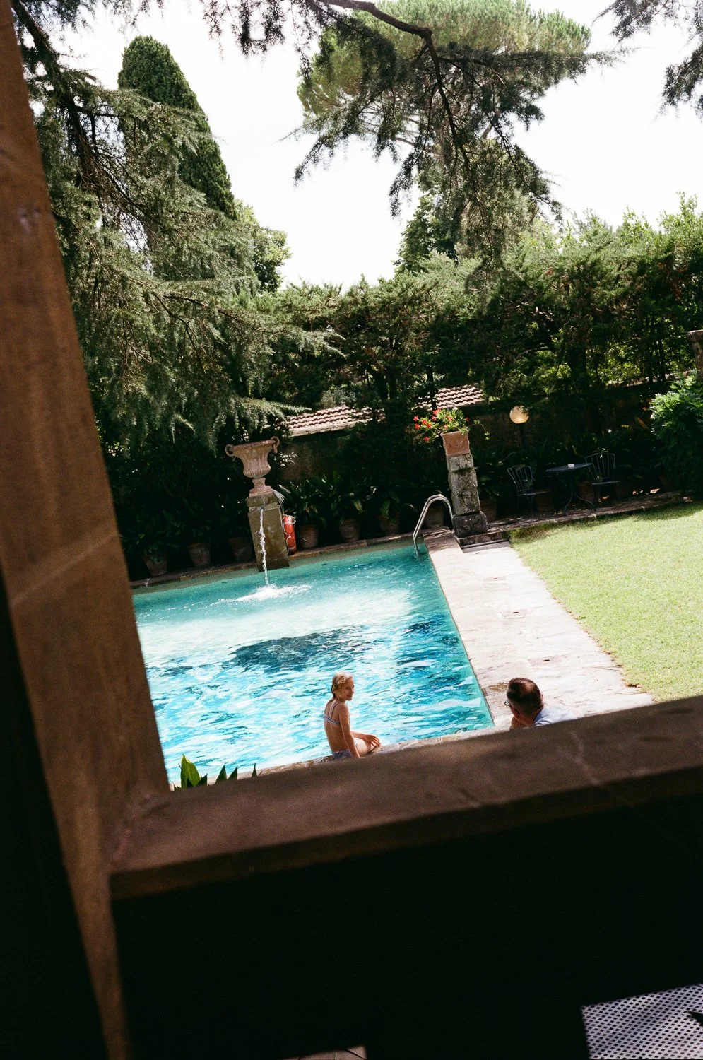 Vista di una piscina all'aperto circondata da alberi e piante, con due persone che si rilassano vicino all'acqua, una ragazza seduta sul bordo e un uomo che la guarda.