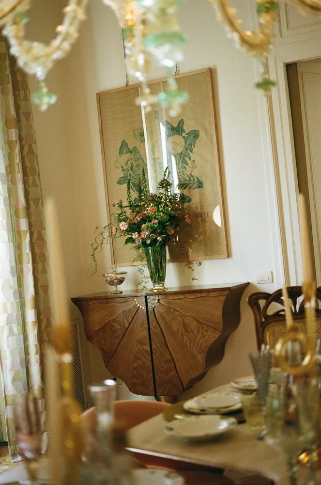 Un'ampia sala da pranzo decorata elegantemente con un grande vaso di fiori colorati su una sideboard in legno, con un quadro con foglie dietro e una tavola apparecchiata con piatti e bicchieri in primo piano.