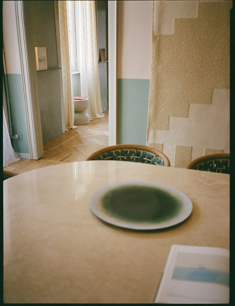 Una stanza con un tavolo di legno, due sedie, un piatto su di esso e una parete con tappezzeria a motivi geometrici. In fondo, una porta verso un'altra stanza con tende chiare, un'alcova con mensole e un water.