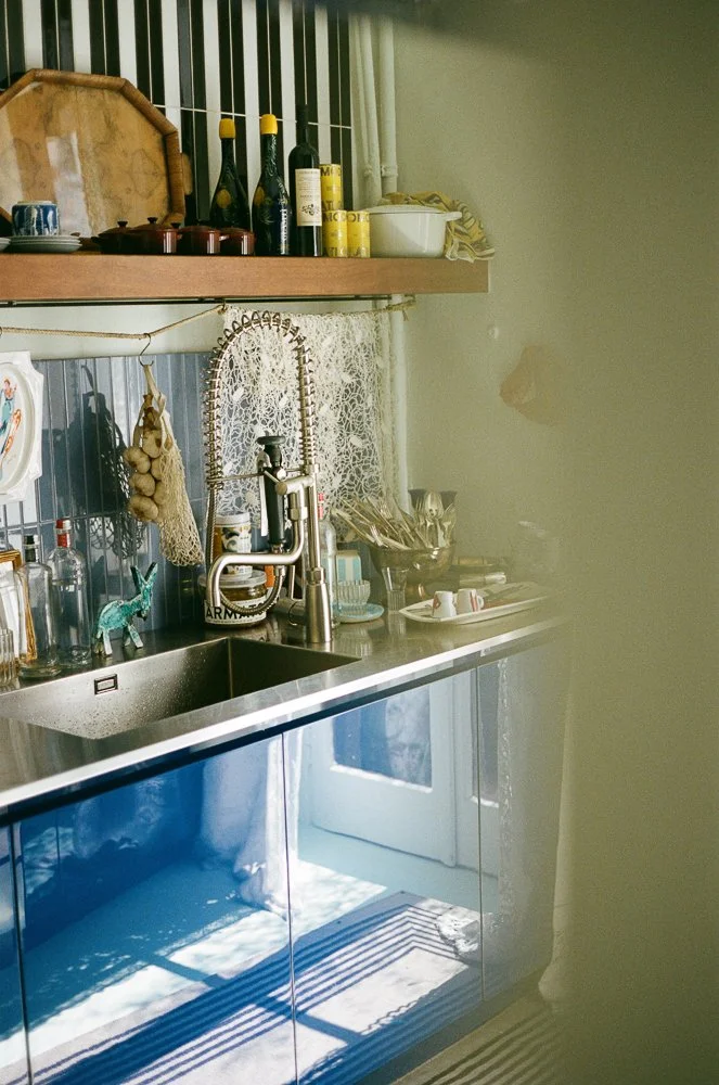 Angolo di una cucina con lavabo in acciaio, sopra uno scaffale con bottiglie di vino, barattoli e altri oggetti. Sul lavandino ci sono piatti, tazze e utensili vari, con uno sfondo di tende ricamate e una porta finestra che lascia entrare luce natura