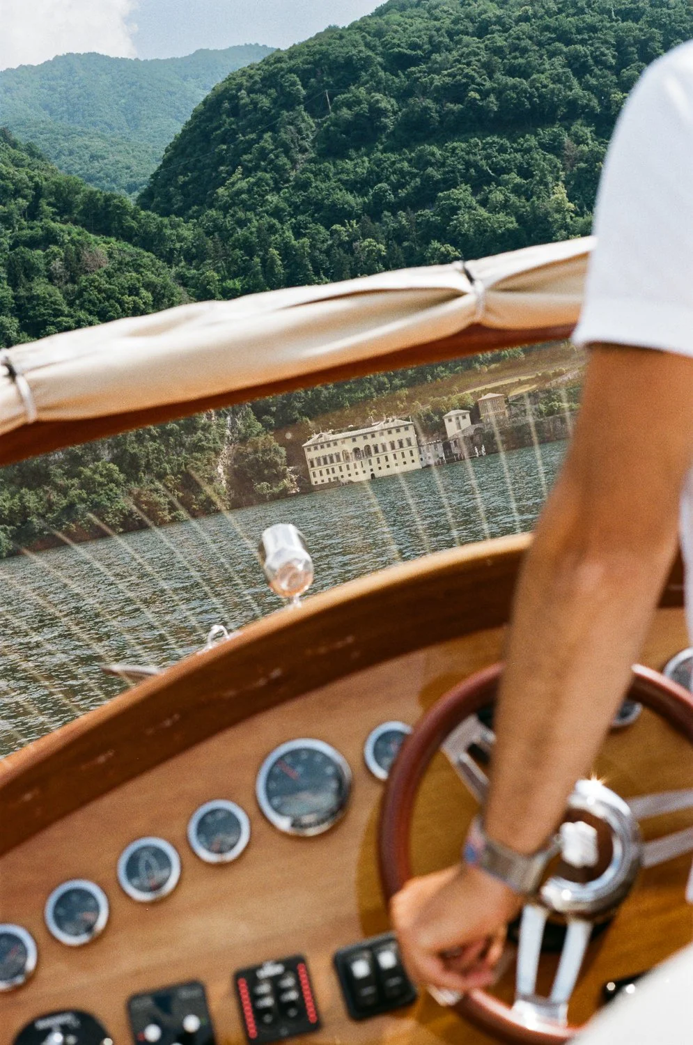 Vista di una barca che naviga su un lago, con uno sfondo di colline verdi e un edificio bianco sulla riva.