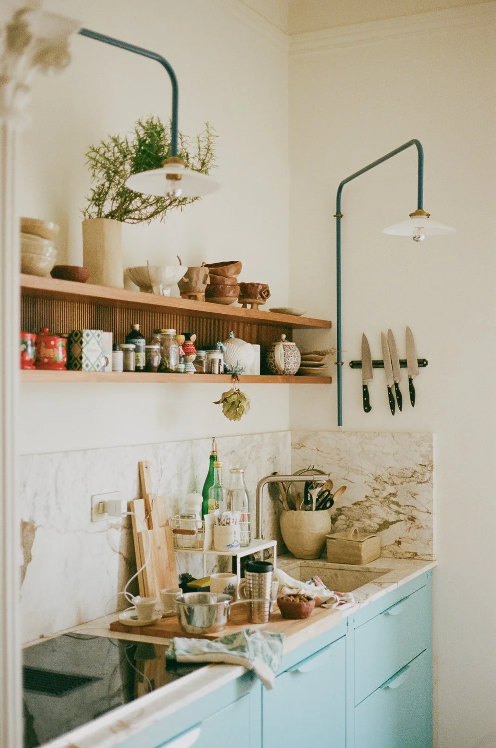 Una cucina con mensole di legno, utensili da cucina, piante, barattoli e utensili vari, con un mobile azzurro e pareti di marmo.