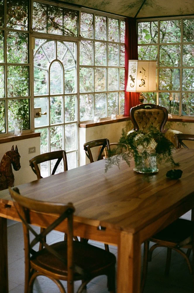 Interior di una stanza da pranzo con grandi finestre che mostrano un giardino verde fuori. Al centro c'è un tavolo in legno con un vaso di fiori davanti a una sedia imbottita con schienale alto. Ci sono anche alcune sedie di legno attorno al tavolo e