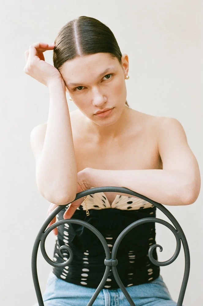 Ragazza con capelli castani, in posa con braccia appoggiate su una sedia in ferro battuto, indossa un top nero con dettagli beige e jeans azzurri, sfondo neutro.